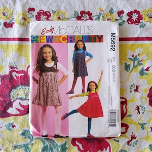 May include: A sewing pattern package featuring three young girls modeling dresses. The package is labeled "Easy McCall's Sew & Crafty" with the pattern number M5692. The size range is CDD (2-3-4-5). The background is a floral patterned fabric.