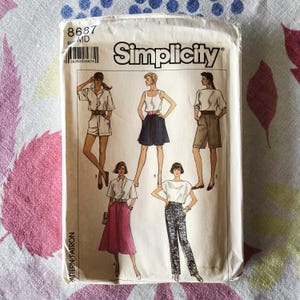 May include: A vintage Simplicity sewing pattern packet, featuring illustrations of various women's clothing designs. The packet is white with the number "8687" and the brand name "Simplicity" printed on it. The designs include shorts, skirts, and pants.