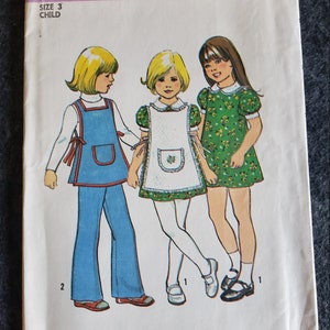 Può includere: Modello di cucito vintage Simplicity numero 7064 per un vestito e pantaloni per bambini. Il modello include tre stili diversi: un vestito floreale verde, un vestito con grembiule bianco e un set di pantaloni e top blu.