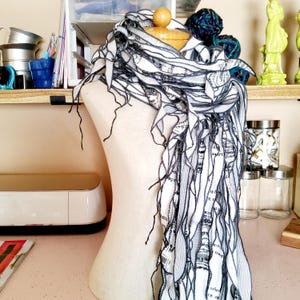 Può includere: Sciarpa bianca con testo nero e disegni a linee, drappeggiata su un manichino. La sciarpa presenta lunghi bordi sfrangiati e un aspetto fluido e a strati. Lo sfondo include mensole con vari oggetti.
