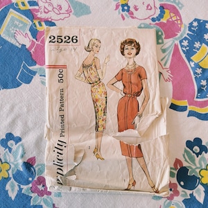 May include: A vintage Simplicity sewing pattern envelope, number 2526, size 14. The paper envelope features illustrations of two dresses, one with a floral print and one in solid orange, with visible creases and tears along the bottom edge.