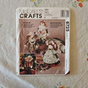 May include: A McCall's Crafts sewing pattern envelope, number 6725, featuring illustrations of three handmade dolls with button accessories. The text states these dolls are 11 inches tall and come complete with clothes and button accessories.