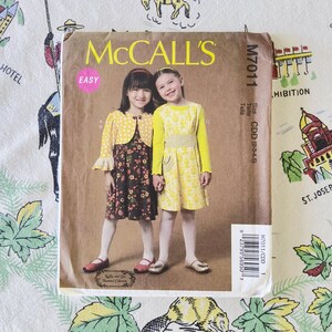 Puede incluir: Un sobre de patrón de costura McCall's M7011 con dos niños vistiendo vestidos. Uno lleva un bolero amarillo de lunares sobre un vestido floral oscuro, y el otro un vestido amarillo de manga larga con estampado. El texto incluye M7011 y CDD (2-3-4-5).