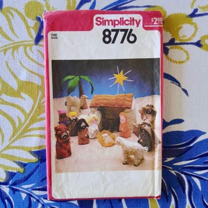 May include: A vintage Simplicity 8776 sewing pattern featuring a nativity scene. The pattern cover is pink and white, with illustrations of the nativity figures, including Mary, Joseph, baby Jesus, and the three wise men. The pattern is one size.