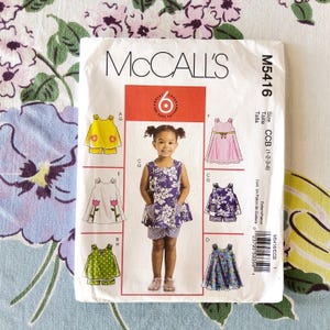 May include: A McCall's sewing pattern envelope, M5416, featuring a young girl and illustrations of children's clothing designs. The designs include dresses and tops with shorts, in various colors and patterns. The envelope is on a floral background.