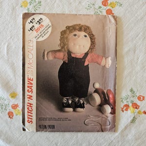 May include: A McCalls Stitch 'N Save sewing pattern envelope for a soft-sculptured doll. The cover features a doll with brown yarn hair, freckles, a red and white checkered shirt, and black overalls. Text includes $1.79 USA, $1.99 CAN, $3.95 AUST, 2015, and fits 16 inch dolls.