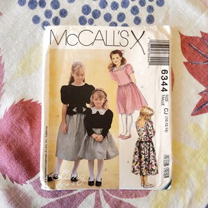 Può includere: Un modello di cucito vintage McCall's per abiti da ragazza. Il modello presenta illustrazioni di ragazze che indossano vari stili di abiti, tra cui un top nero con una gonna a quadretti, un abito rosa e un abito floreale. La taglia del modello è CJ (10,12,14).