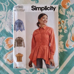 May include: Simplicity sewing pattern for women's tops. The pattern features a woman wearing an orange shirt with a pleated bodice and peplum. The pattern includes illustrations of different top styles and sizing information.
