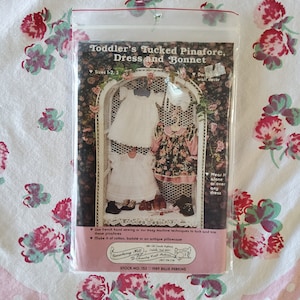 May include: A vintage sewing pattern for a toddler's tucked pinafore dress and bonnet. The pattern is for sizes 1-2 and 3. The pattern is illustrated with three dolls wearing the pinafore dress and bonnet. The pattern is from Strawberry Hill Country Craft Patterns and is stock number 153. The pattern was designed by Billie Perkins.