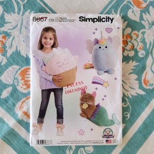May include: Simplicity sewing pattern 8667 featuring plush toy designs. The pattern includes instructions for a cupcake, a cat with wings, and a mermaid cat. The pattern is marked "Excess Discontinued".