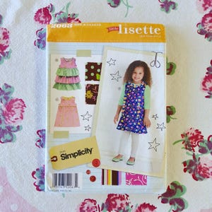 May include: A sewing pattern package featuring a child's dress design. The package has the text "Little Lisette" and "Simplicity". The dress designs include a pink and green tiered dress, a pink polka dot dress, and a blue floral dress. The package is on a floral patterned fabric.