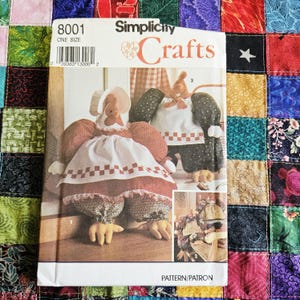 May include: A Simplicity Crafts 8001 sewing pattern envelope featuring illustrations of two fabric chicken dolls. The dolls wear dresses with white aprons, checkered trim, and bonnets. The envelope includes the text 8001, ONE SIZE, and PATTERN/PATRON.