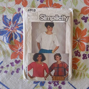 Può includere: Un pacchetto di modelli di cucito Simplicity vintage con tre modelli di camicette. Il pacchetto è bianco con il numero 6913 e il logo Simplicity. I modelli di camicette includono un top bianco, rosso e a quadri.