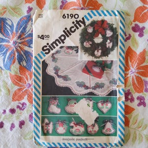 May include: A Simplicity sewing pattern for a Christmas tree skirt and ornaments. The pattern number is 6190. The pattern includes instructions for making a tree skirt with a scalloped edge and a variety of ornaments, including stockings, bells, and hearts. The pattern is printed on a white background with blue and red stripes.