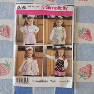 May include: A Simplicity sewing pattern envelope, number 2699, size A 1/2, 1, 2, 3, 4. The front displays four variations of a sleeveless jumper or apron dress for children, featuring different fabric patterns, colors, and decorative details.