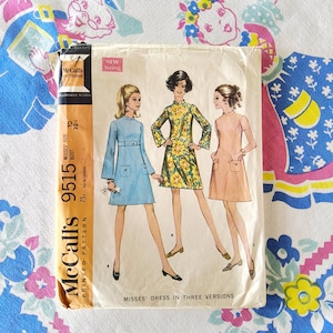 Könnte beinhalten: Vintage McCall's 9515 Schnittmuster für Damenkleider in drei Ausführungen. Das Schnittmuster zeigt Illustrationen von Kleidern in Blau, gelbem Blumenmuster und Rosa. Brustumfang 32 Zoll (81 cm).