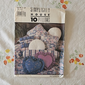 Può includere: Una busta per cartamodello Simplicity House per 10 cuscini, con una fotografia di vari cuscini decorativi, inclusi stili a forma di cuore, rotondi e rettangolari in tessuti floreali e a righe con volant e dettagli trapuntati.