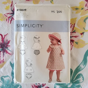 May include: Simplicity sewing pattern number R10609 for a girl's dress and bloomers with a hat. The pattern includes four different styles, with a floral print dress and hat, a solid white dress and hat, and two variations of a dress with a pocket and a hat.