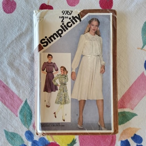 May include: A Simplicity sewing pattern envelope 9767 for a dress in size 14. The cover features illustrations of three dress variations: a solid cream dress with lace detail, a solid purple dress, and a floral print dress with long sleeves.
