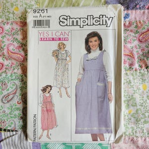 以下が含まれることがあります： Simplicityの型紙封筒、番号9261、サイズA (PT-MD)、タイトルは「Yes I Can Learn to Sew」。白い紙の封筒には、ポケットとリボンのディテールが付いたノースリーブのジャンパースカートの3つのバリエーションのイラストが描かれています。