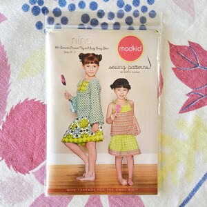 May include: A sewing pattern package with two children modelling clothes. The package reads "nina" and "All-Season's Peasant Top and Easy Peasy Shirt" with sizes 2-7. The "modkid" logo is visible, along with "sewing patterns" and "MOD THREADS FOR THE COOL KID".