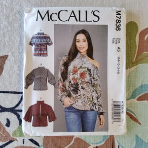 May include: McCall's sewing pattern M7836. The pattern features illustrations of tops with various sleeve and neckline designs. The package includes a photo of a woman wearing a floral print top. Size A5 (6-8-10-12-14) is indicated on the package.