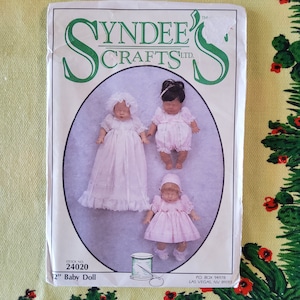 May include: A vintage sewing pattern for a 12-inch baby doll. The pattern is from SynDee's Crafts LTD and features three dolls in pink dresses. The pattern number is 24020.