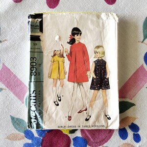 Puede incluir: Sobre de patrón McCall's vintage con ilustraciones de vestidos de niña en tres versiones. El sobre está desgastado, con una franja verde y negra en el lateral. Las ilustraciones muestran a niñas con vestidos rojos, amarillos y negros. El texto dice "GIRLS' DRESS IN THREE VERSIONS."