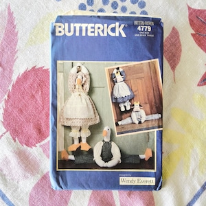 Puede incluir: Un sobre de patrón Butterick azul con un diseño de muñeca de ganso. El sobre muestra una muñeca de ganso terminada con un vestido y un delantal, con diseños de muñecas de vaca adicionales. El texto "Designed by Wendy Everett" es visible.