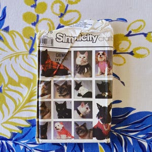 May include: A vintage Simplicity Crafts pattern for cat and dog apparel. The pattern features images of various cats and dogs in different outfits. The packaging is white with the Simplicity logo and the words "Catwear Dogwear". The background is a floral pattern.