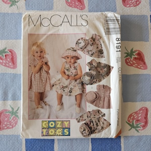 May include: A McCalls 8191 sewing pattern envelope for children's clothing titled Cozy Togs. The front features photographs of children wearing dresses and hats, along with illustrations of various dress and hat designs in floral and gingham prints.