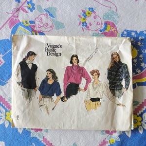 Peut inclure: Une pochette de patron de couture Vogue's Basic Design présentant des illustrations de cinq hauts différents étiquetés de A à E. L'enveloppe en papier blanc affiche divers styles, dont un gilet, un chemisier bleu, un haut rose et des chemises à carreaux.