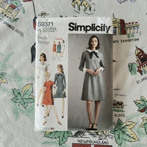 Op de afbeelding: Een Simplicity naaipatroon envelop S9371 voor een jurk in 1960s vintage stijl. De voorzijde toont illustraties van drie jurkvariaties en een foto van een grijze jurk met een witte kraag, witte manchetten en een zwarte strik bij de hals.