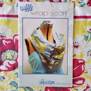 May include: A printed pattern for a ruffle wrap scarf. The scarf is displayed on a dress form, showcasing a blue, yellow, and white patterned fabric. The text "ruffle wrap scarf" is at the top, with "skburton designs" at the bottom.