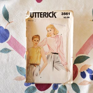 May include: A vintage Butterick sewing pattern, size 10, featuring illustrations of two women's blouses. The pattern cover has the number 3561 and the price $2.00. The background is a floral patterned fabric.