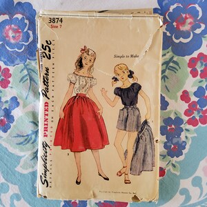 May include: Vintage Simplicity sewing pattern number 3874, size 7, for a skirt and shorts. The pattern is printed on light brown paper with a white border. The pattern includes instructions for making a red skirt and blue and white striped shorts.