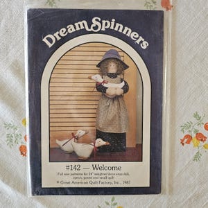 Puede incluir: Un folleto de patrones Dream Spinners #142 titulado Welcome, que incluye una muñeca tope de puerta de 61 cm, delantal, ganso y edredón. La portada muestra una muñeca con sombrero azul y vestido oscuro sosteniendo un ganso blanco y dos gansos.
