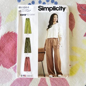May include: Simplicity sewing pattern R11944 for wide-leg pants. The pattern includes illustrations of the pants in green, patterned black, and red. The model in the image is wearing a white sweater and brown pants. The pattern is easy-to-sew.