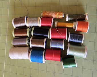 Puede incluir: Una colección de carretes de hilo en varios colores, incluyendo marrón, plateado, rojo, negro, azul y verde. Los carretes están dispuestos sobre una esterilla de corte verde, mostrando una variedad de suministros de costura y artesanía.