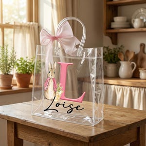 May include: Transparent tote bag with a pink bow and handles. The bag displays a pink letter "L" and a watercolour illustration of a rabbit in a pink jacket. The name "Loize" is written in cursive below the letter.