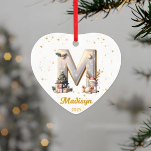 May include: Heart-shaped Christmas ornament with the letter "M" in silver and gold. The ornament features a Christmas tree, a reindeer wearing a Santa hat, and the name "Madisyn" with the year "2025". A red ribbon is attached for hanging.