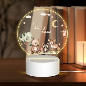Puede incluir: Luz de noche personalizada con una escena del bosque: oso, mapache, ciervo y búho. El panel acrílico está iluminado por una base blanca, con el nombre "Jenny" y la fecha "10.09.2025". El diseño incluye un árbol, nubes, estrellas y una luna creciente.