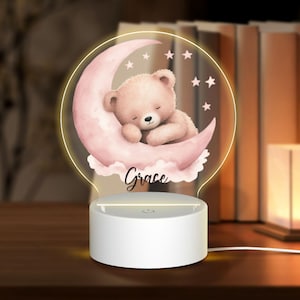 Könnte beinhalten: Nachtlicht mit einem schlafenden Teddybär auf einem rosa Mond und Sternen. Der Name "Grace" steht darunter. Das Licht hat einen weißen Sockel und einen warmen Schein.