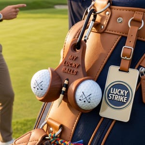 Könnte beinhalten: Eine Golftasche mit braunen Lederakzenten und blauem Innenraum. Zwei Golfbälle werden in einem braunen Halter mit der Aufschrift "MY LUCKY BALLS" gehalten. Ein Anhänger mit der Aufschrift "LUCKY STRIKE" hängt an der Tasche.