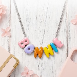 Pode incluir: Um colar colorido que soletra "BONHINI" em contas de letras de bolhas coloridas em tons pastel. O colar tem uma corrente prateada e está sobre um fundo de madeira branca com flores e acessórios cor-de-rosa.