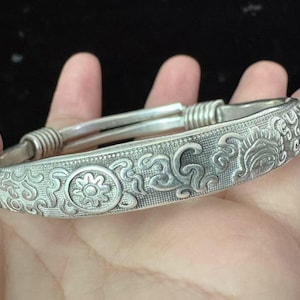 May include: A silver-colored bangle bracelet with intricate floral and scrollwork designs. The bracelet features a central floral motif and detailed patterns along its surface. The ends of the bracelet have a coiled design.