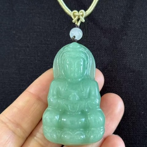 May include: A jade pendant featuring a seated Buddha figure in a light green hue. The pendant is suspended from a gold-toned cord with a knot detail and a small white bead. The Buddha is carved with detailed features.