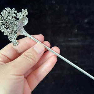 May include: A silver-toned hair stick with a floral design. The decorative top features detailed flowers and leaves, with a curved, ornate accent. The stick is held against a dark background.