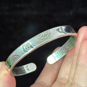 May include: A silver cuff bracelet with etched designs and symbols. The open-ended bracelet features decorative patterns and what appears to be Asian characters. The bracelet is held in a hand against a dark background.