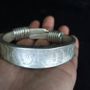 Könnte beinhalten: Silberfarbenes Armband mit aufwendigen eingravierten Mustern. Das Armband hat ein zentrales Design und dekorative Elemente auf dem Band. Die Enden sind mit gewickeltem Silberdraht verbunden. Ein klassisches Schmuckstück.